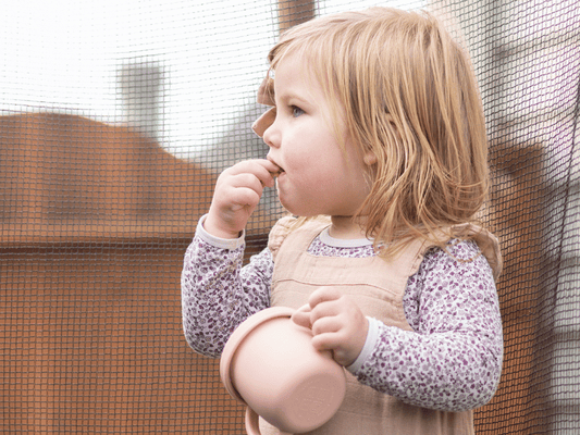 Toddler girl holding a silicone cup and biting her fingers while standing outdoors, showcasing the practical use of child-safe silicone products.