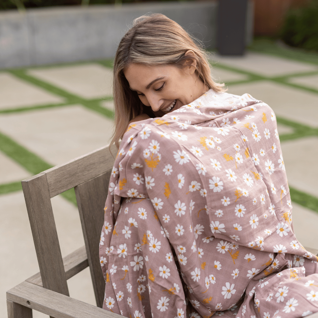 Woman wrapped in a daisy-print bamboo swaddle, smiling while seated outdoors.