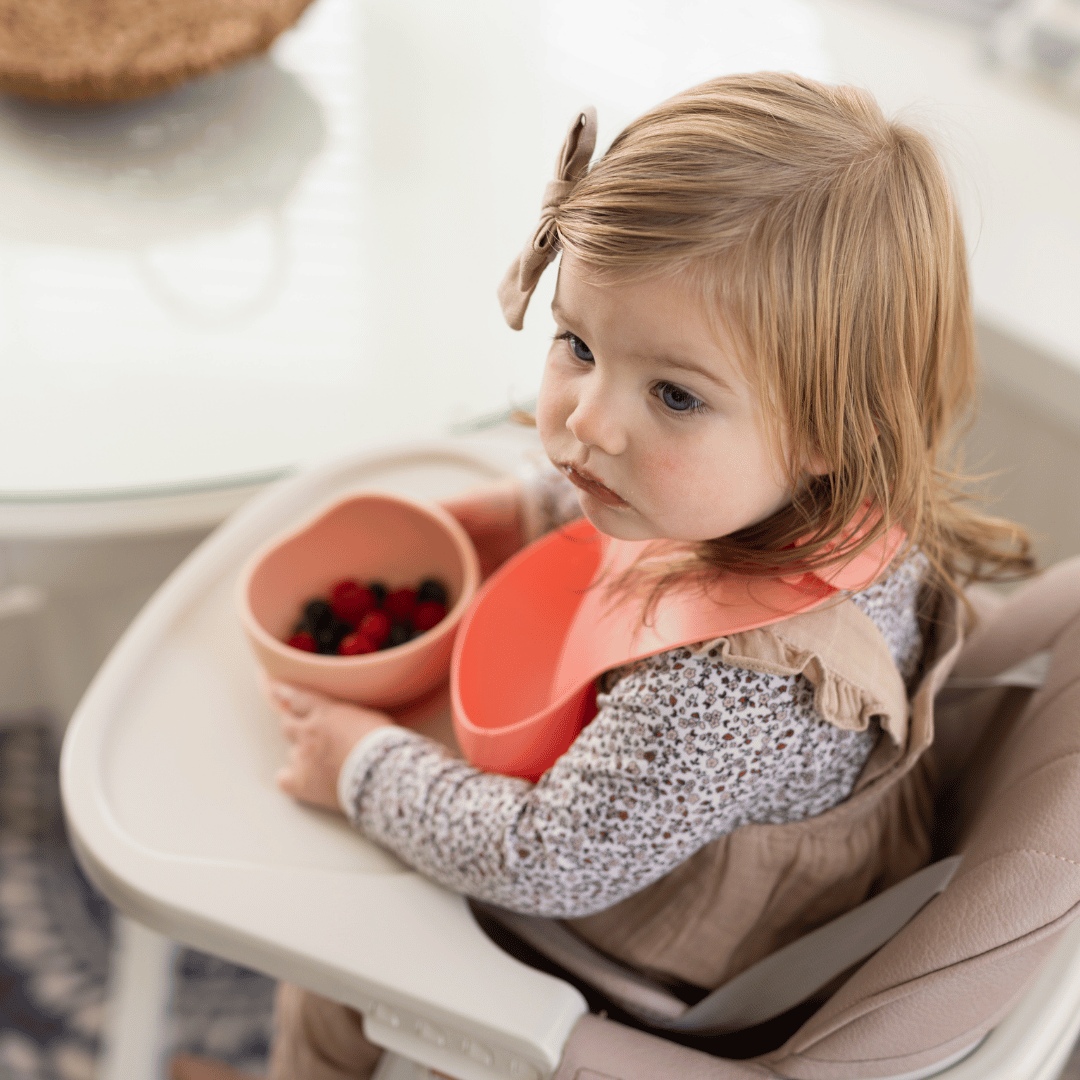 Toddler girl sitting in a high chair, eating berries from pink silicone bowls, reflecting a mess-free feeding solution.