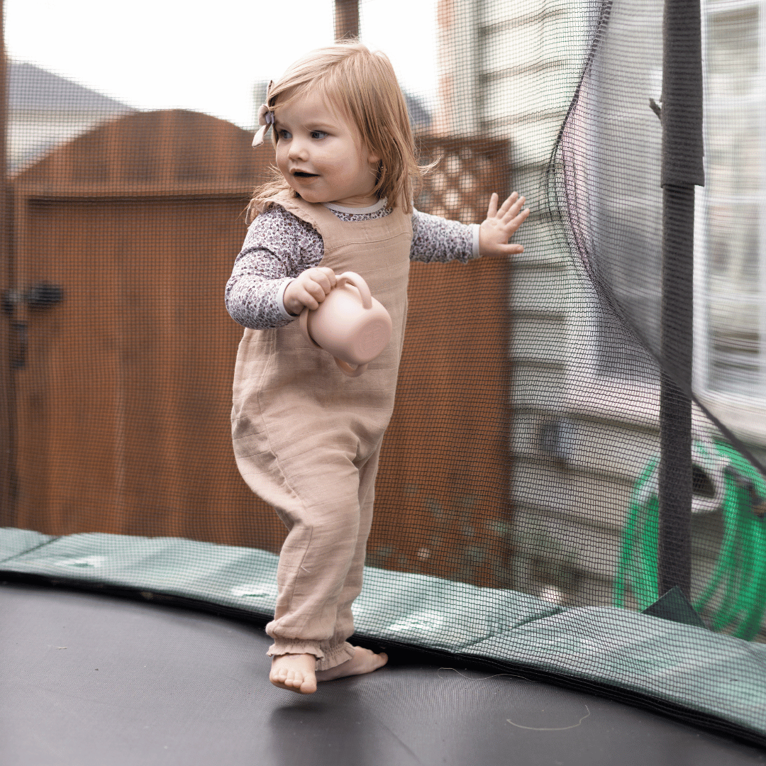 Active toddler in overalls holding a silicone snack cup while standing on a trampoline, surrounded by safety netting.