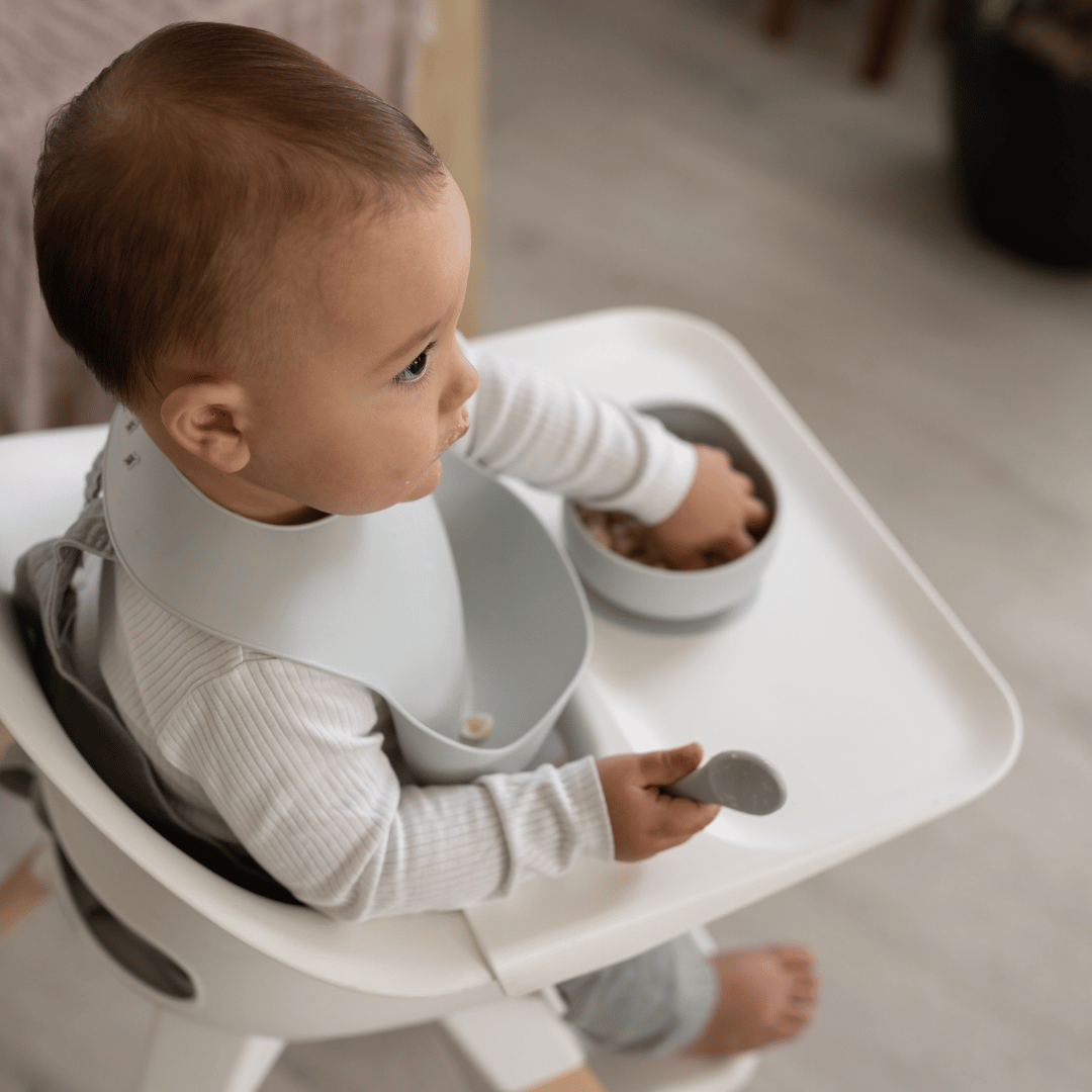 Baby in a highchair wearing a cloud-colored silicone bib, holding a silicone spoon and reaching into a silicone bowl filled with cereal.