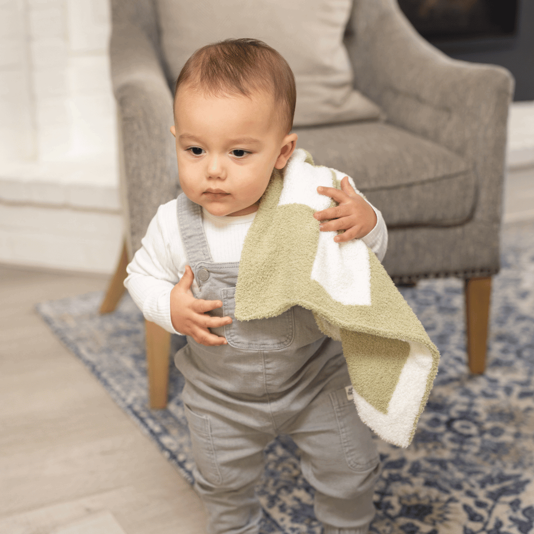 Toddler holding a Luxe Checkered Mini Baby Blanket in Matcha Green, seated near a cozy living room chair, looking thoughtful.