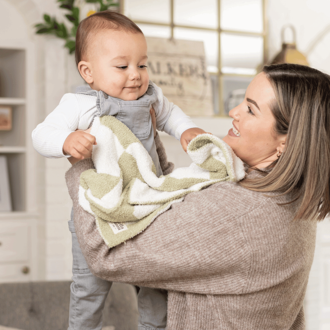 Mother smiling at her baby who is holding a Luxe Checkered Mini Baby Blanket in Matcha Green, capturing a tender family moment at home.