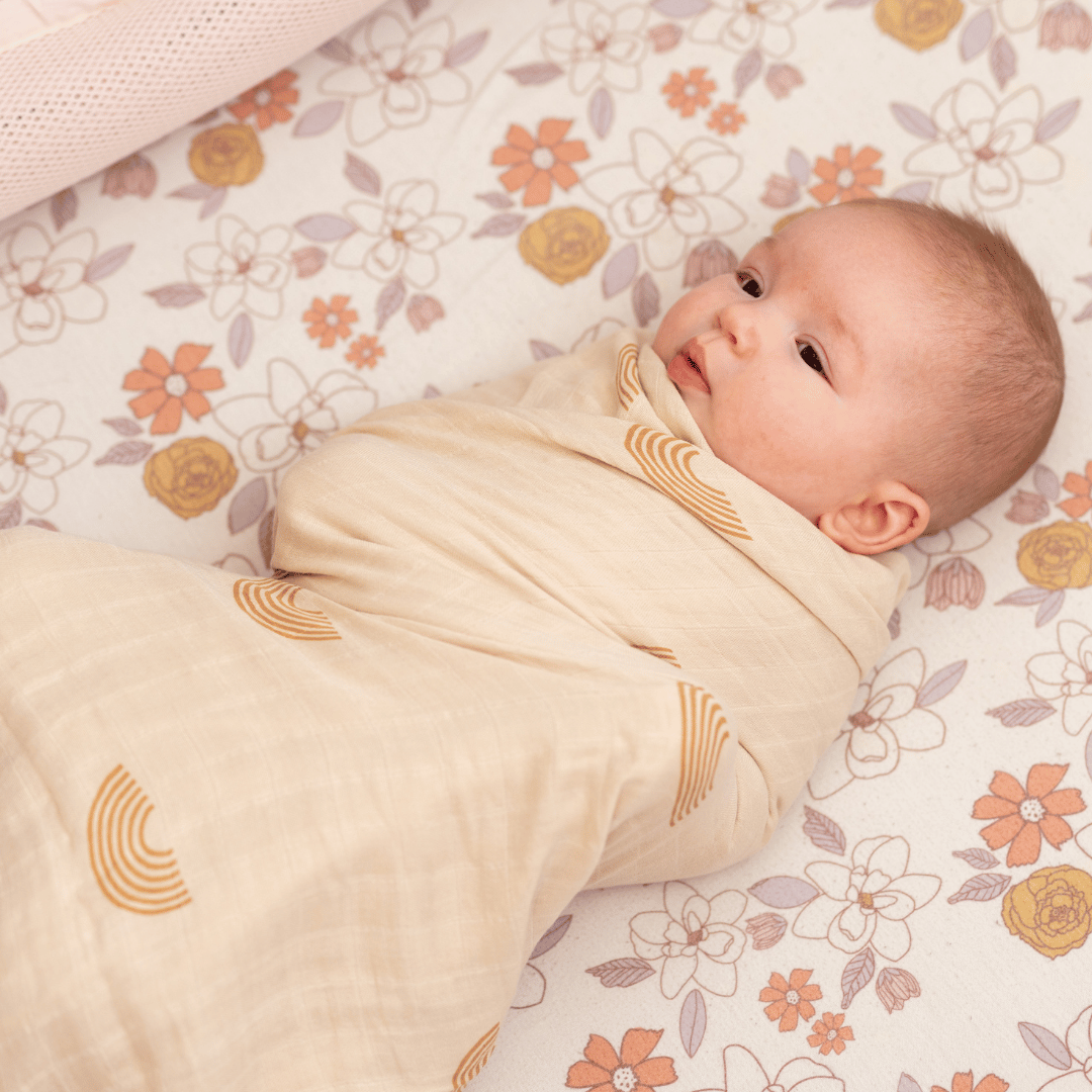 Baby swaddled in a soft beige bamboo blanket with rainbow prints, lying on a floral-patterned crib sheet.