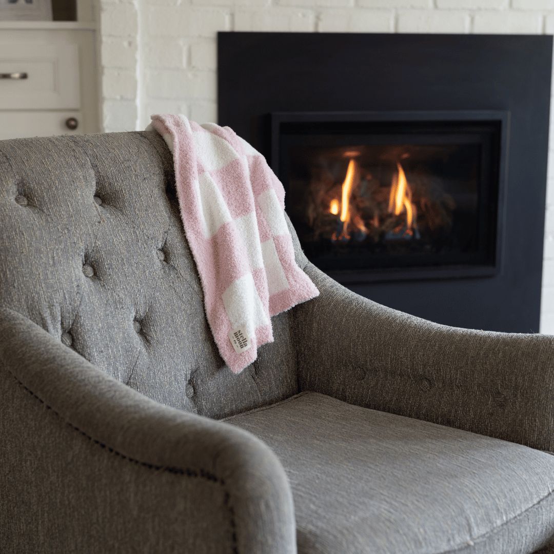 Pink and white luxe checkered baby blanket draped over a gray armchair near a cozy fireplace.