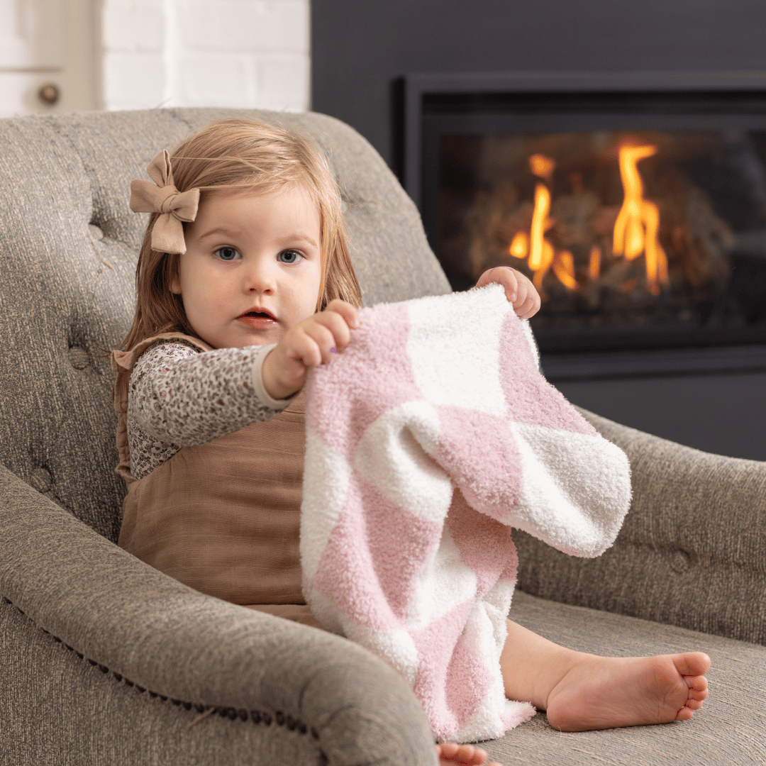  Little girl holding a pink and white luxe checkered baby blanket, sitting on a cozy chair in front of a warm fireplace.