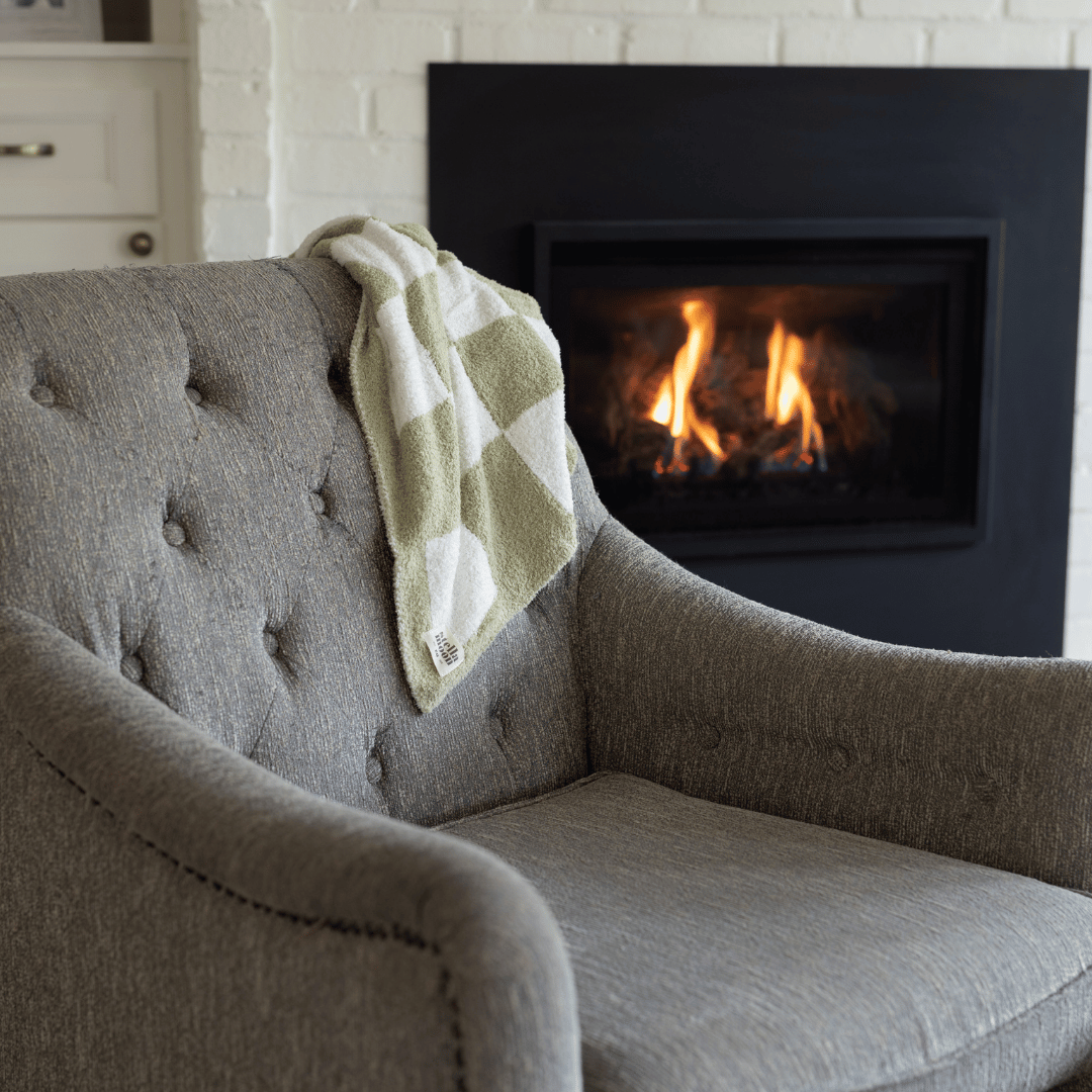 Matcha Luxe Blanket draped over a gray tufted chair, featuring a soft green and white checkered design, with a cozy fireplace in the background.