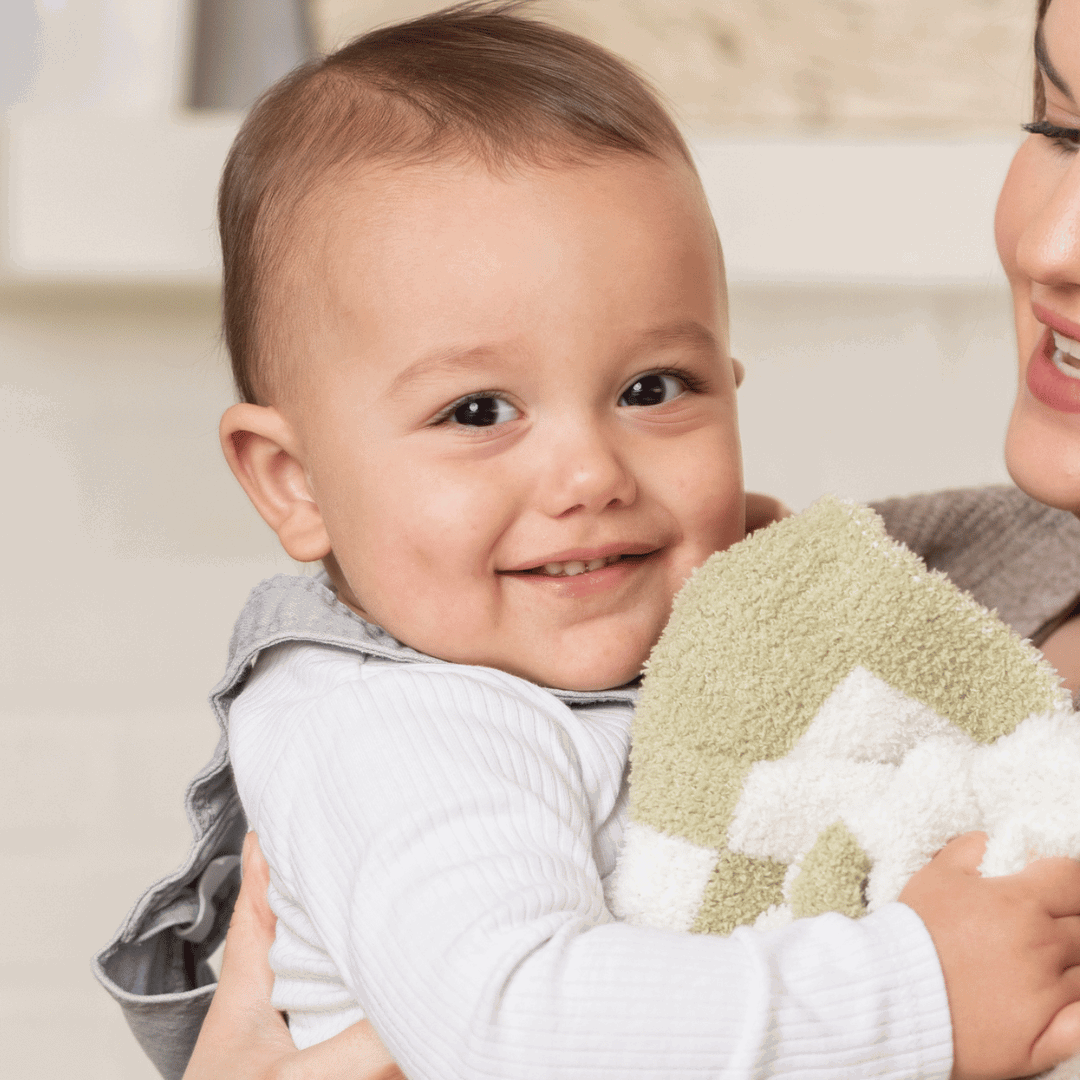 Smiling baby held closely by mother, both enjoying a warm moment with a Luxe Checkered Mini Baby Blanket in Matcha Green in the background.