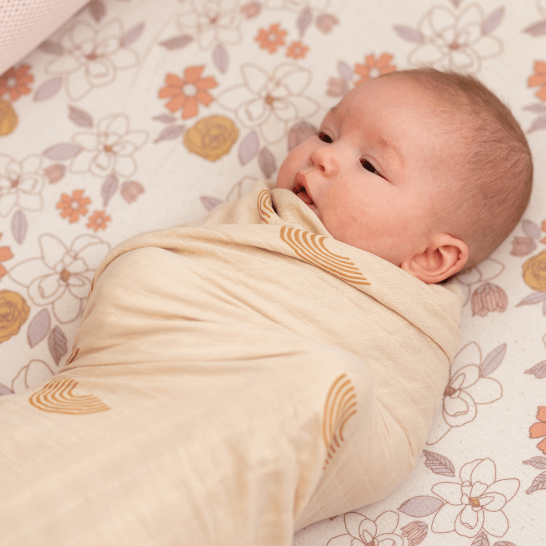 Newborn baby snugly wrapped in an organic bamboo swaddle blanket with a rainbow design, lying on a floral-patterned crib sheet.