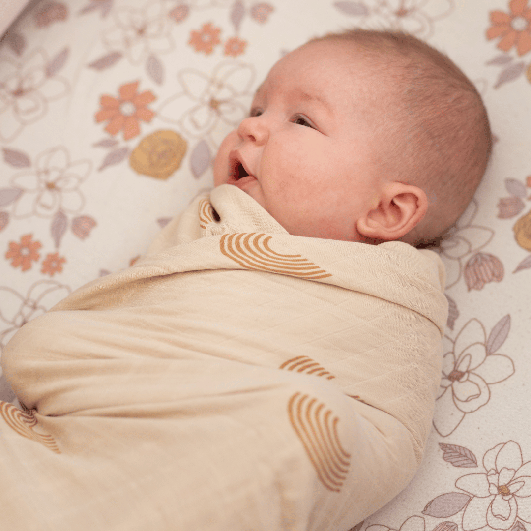 A newborn baby wrapped in a soft organic bamboo swaddle blanket with a minimalist rainbow pattern, lying on a floral crib sheet.