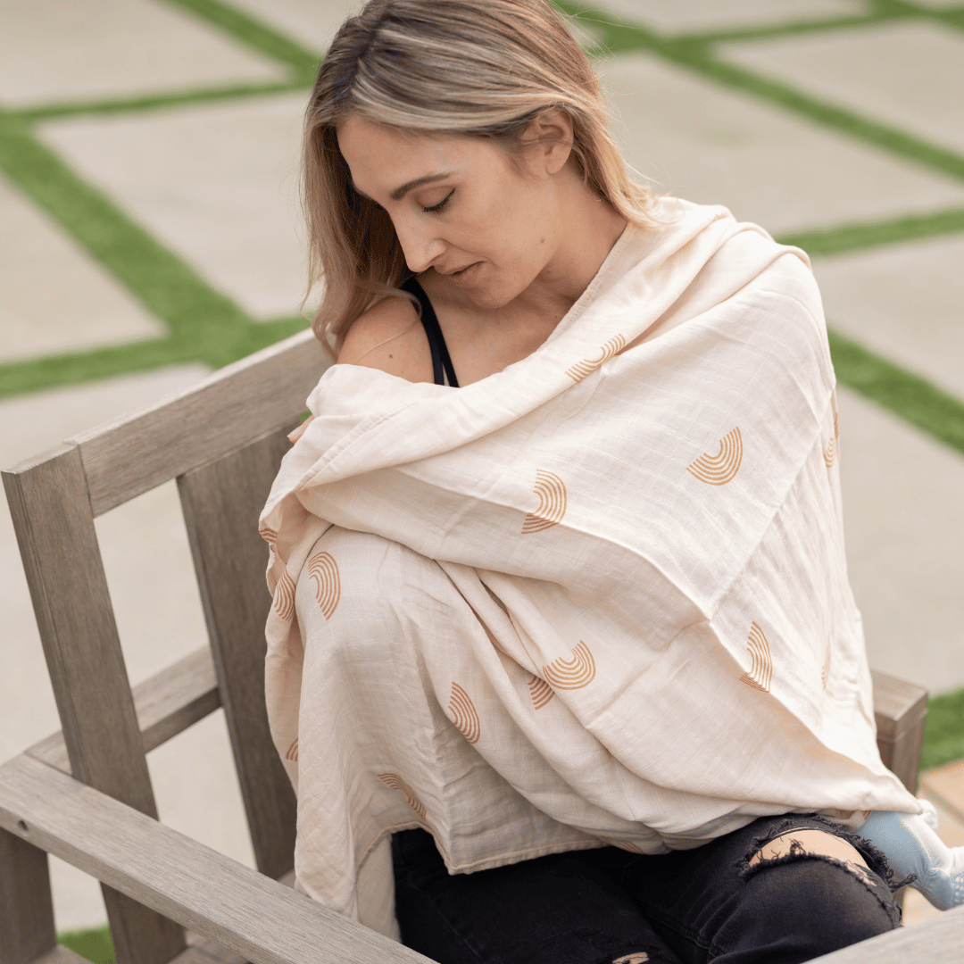 A mother using an organic bamboo swaddle blanket with a minimalist rainbow pattern as a breathable and lightweight breastfeeding cover outdoors.