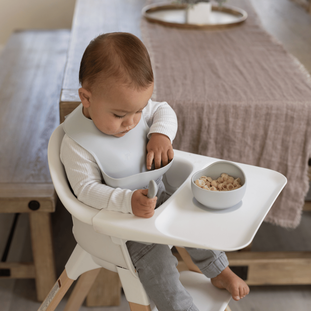 Baby in a highchair wearing a cloud-colored silicone bib, using a silicone spoon to eat cereal from a matching bowl.