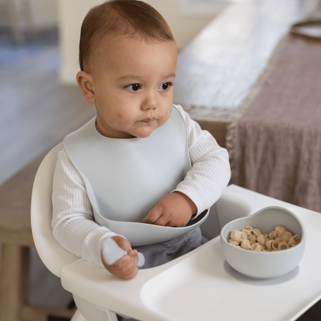 Baby wearing a cloud-colored silicone bib, holding a silicone spoon while eating cereal from a heart-shaped silicone bowl in a highchair.