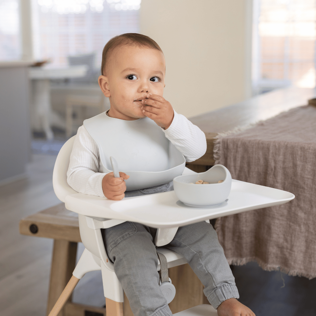 Baby wearing a cloud-colored silicone bib while sitting in a highchair, eating cereal from a matching silicone bowl.