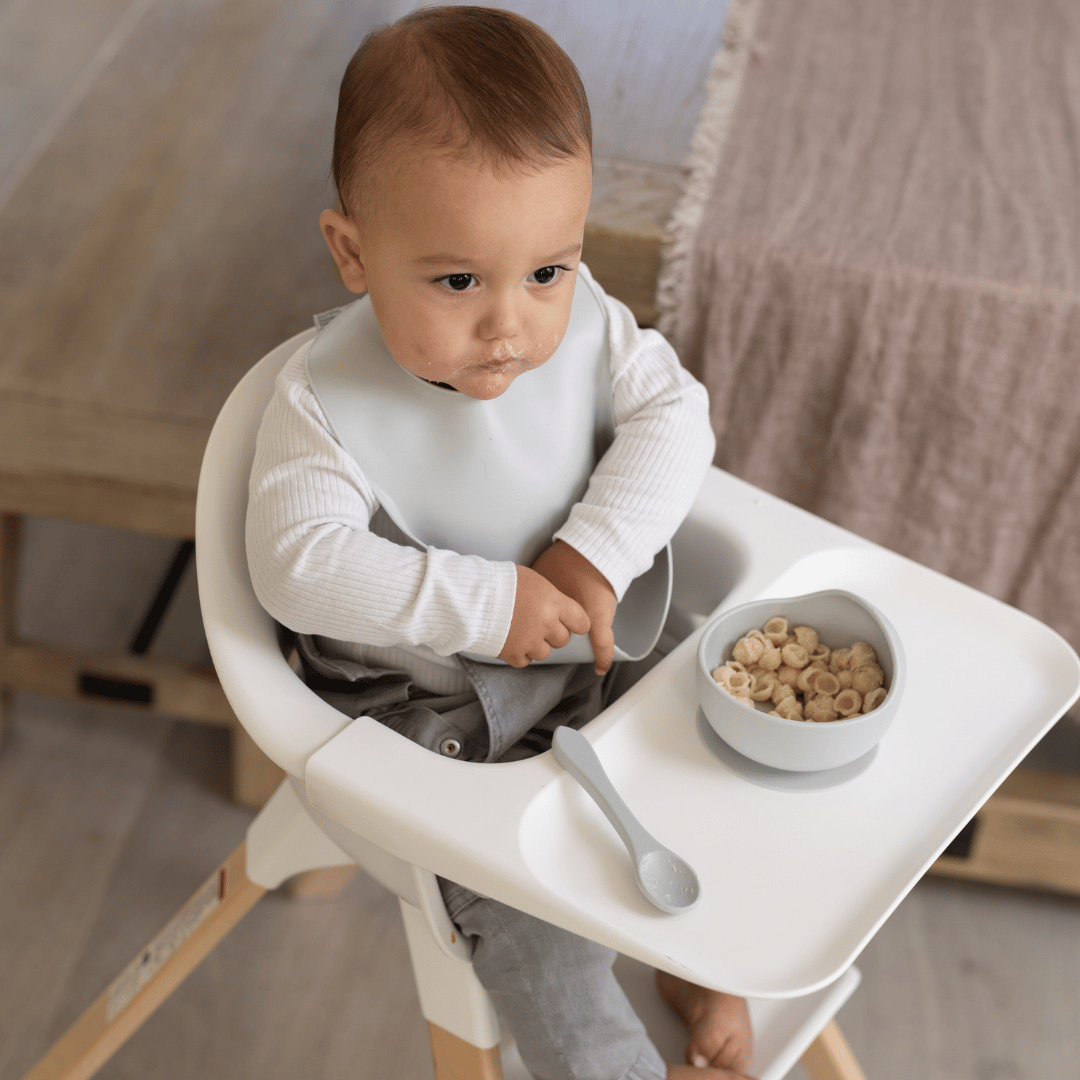 Baby in a high chair wearing a silicone bib, using a Silicone Suction Bowl & Spoon Set in Cloud with pasta. Ideal for mess-free, BPA-free baby feeding.