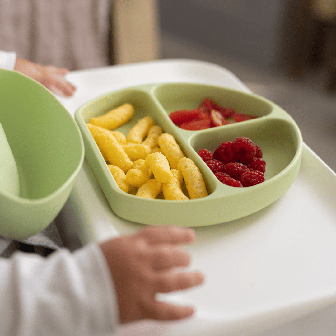 Close-up of a silicone suction plate with divided sections containing snacks—puffed crisps, raspberries, and sliced tomatoes—on a baby's high chair tray.