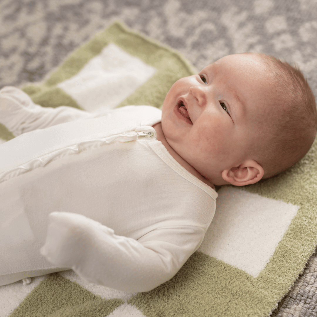 Joyful baby smiling while lying on a Luxe Checkered Mini Baby Blanket in Matcha Green by Stella Moon Co., enhancing a moment of comfort and style.