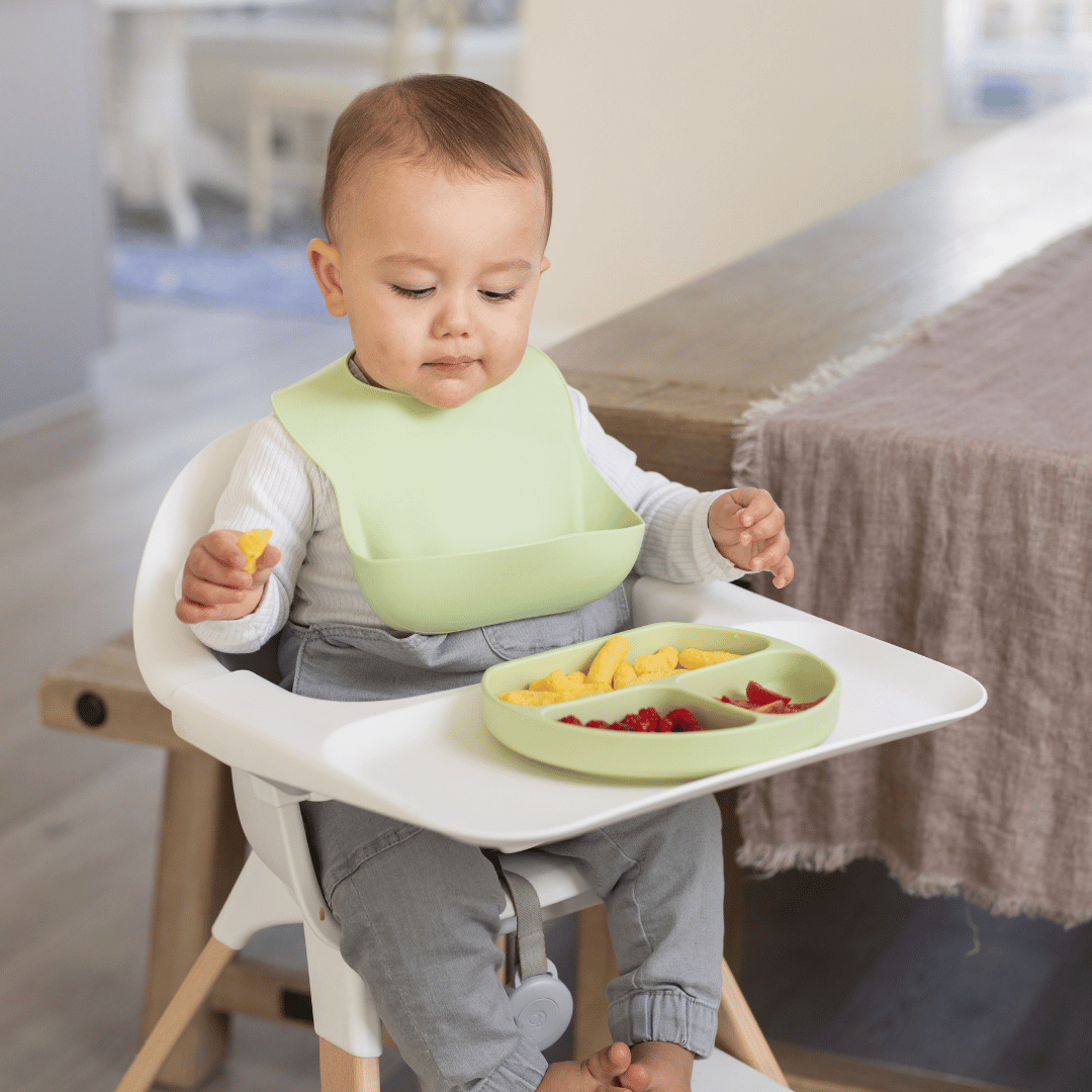 Baby wearing a spearmint silicone bib, eating snacks from a divided suction plate in a high chair.