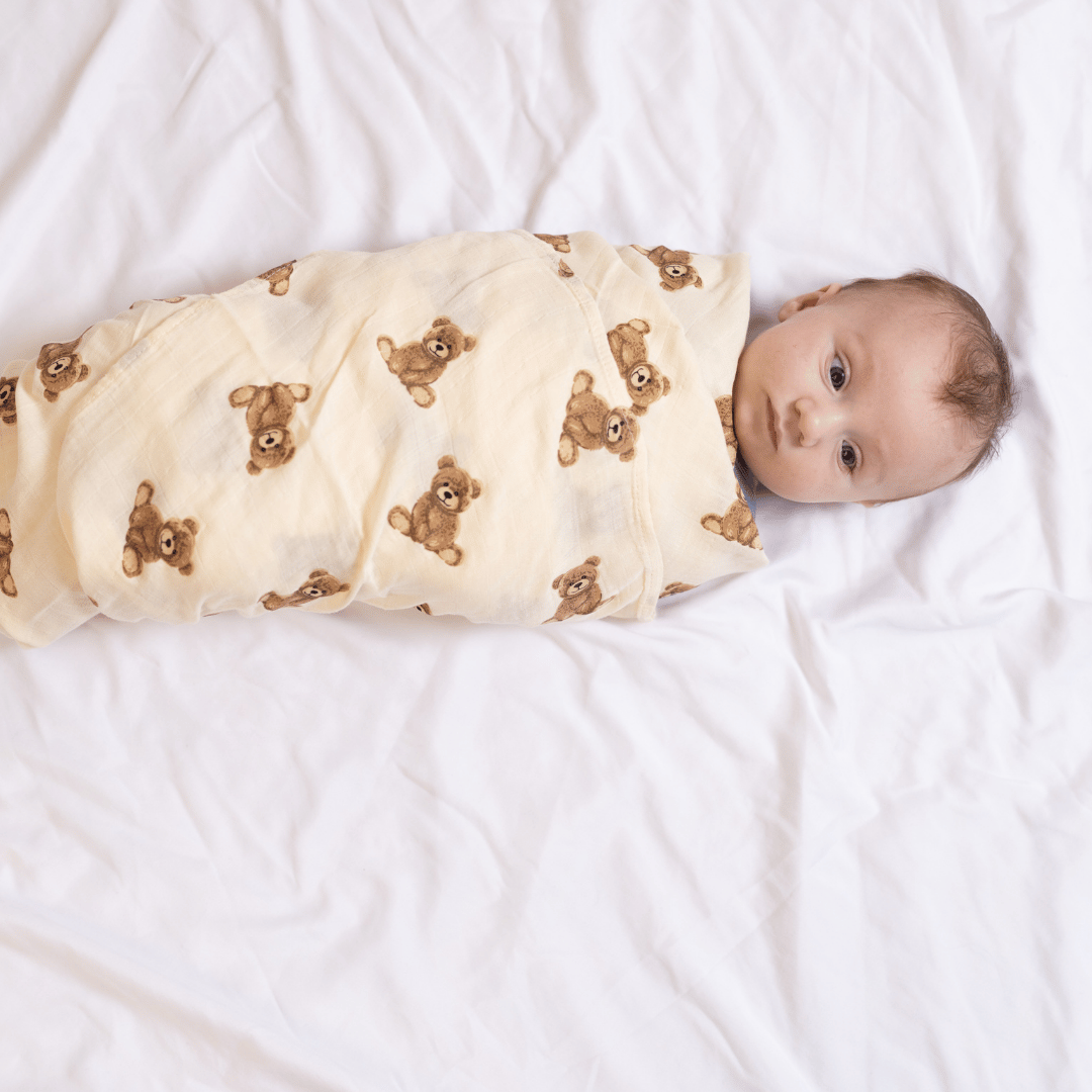 Baby swaddled snugly in the Stella Moon Multi-Use Bamboo Swaddle in Teddy. The soft bamboo-cotton blend blanket features an adorable teddy bear print, providing warmth and comfort on a cozy white bed.