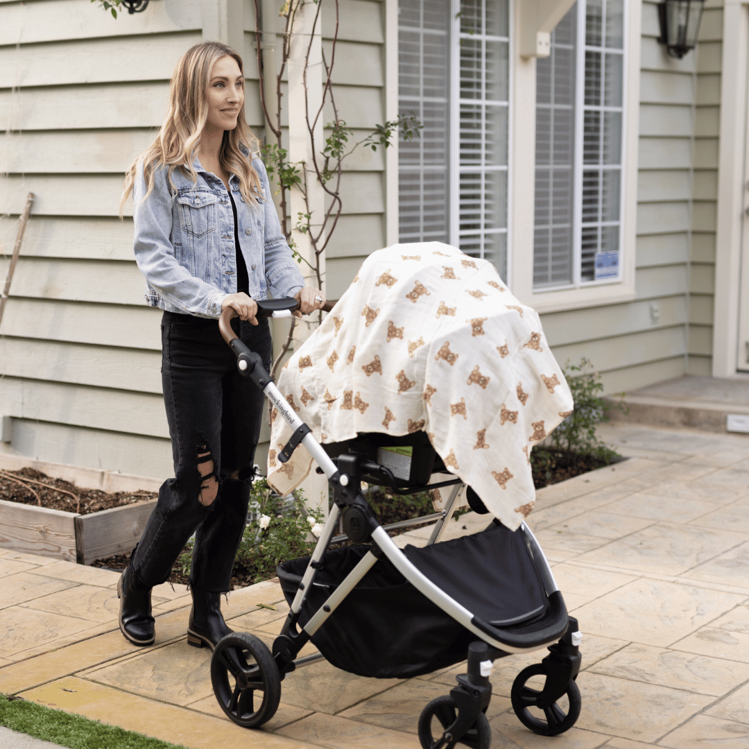 Mother pushing a stroller covered with the Stella Moon Multi-Use Bamboo Swaddle in Teddy. The soft, breathable swaddle features an adorable teddy bear print, providing shade and comfort for the baby.