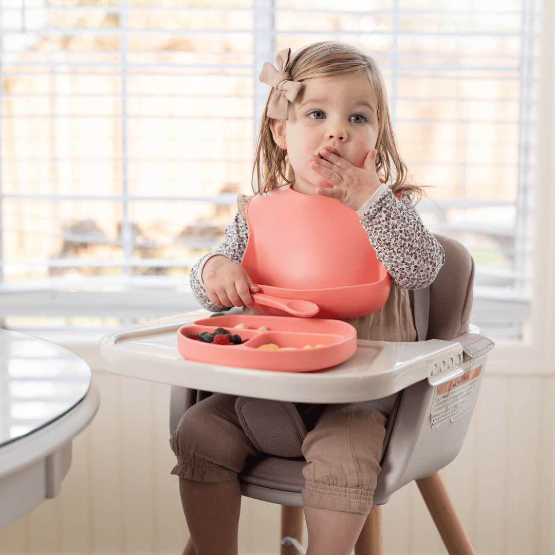 Toddler in a high chair enjoying a meal with the Stella Moon silicone suction plate in Sherbert. The child is wearing a matching silicone bib with a food catcher and eating independently, with a divided plate holding fresh fruit and snacks.