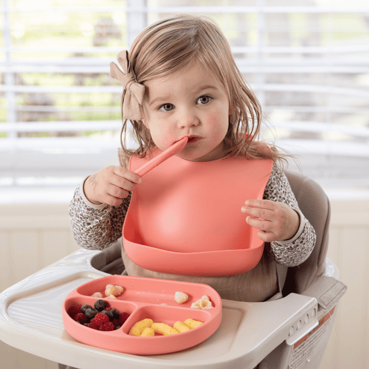 A toddler sitting in a high chair using the Stella Moon silicone suction plate in Sherbert. The child is wearing a matching silicone bib with a food catcher and holding a spoon while eating a balanced meal. The divided plate contains fresh fruit, cheese, and snacks, designed for mess-free self-feeding.