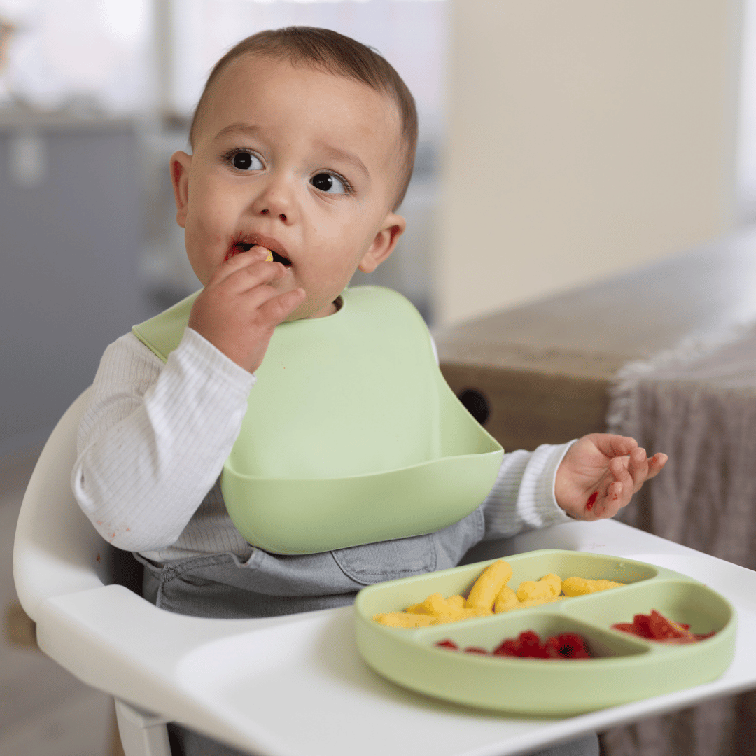 Baby sitting in a high chair using the Stella Moon silicone suction plate in Spearmint. The baby is wearing a matching silicone bib with a food catcher while self-feeding with a divided plate containing fresh berries and snacks.