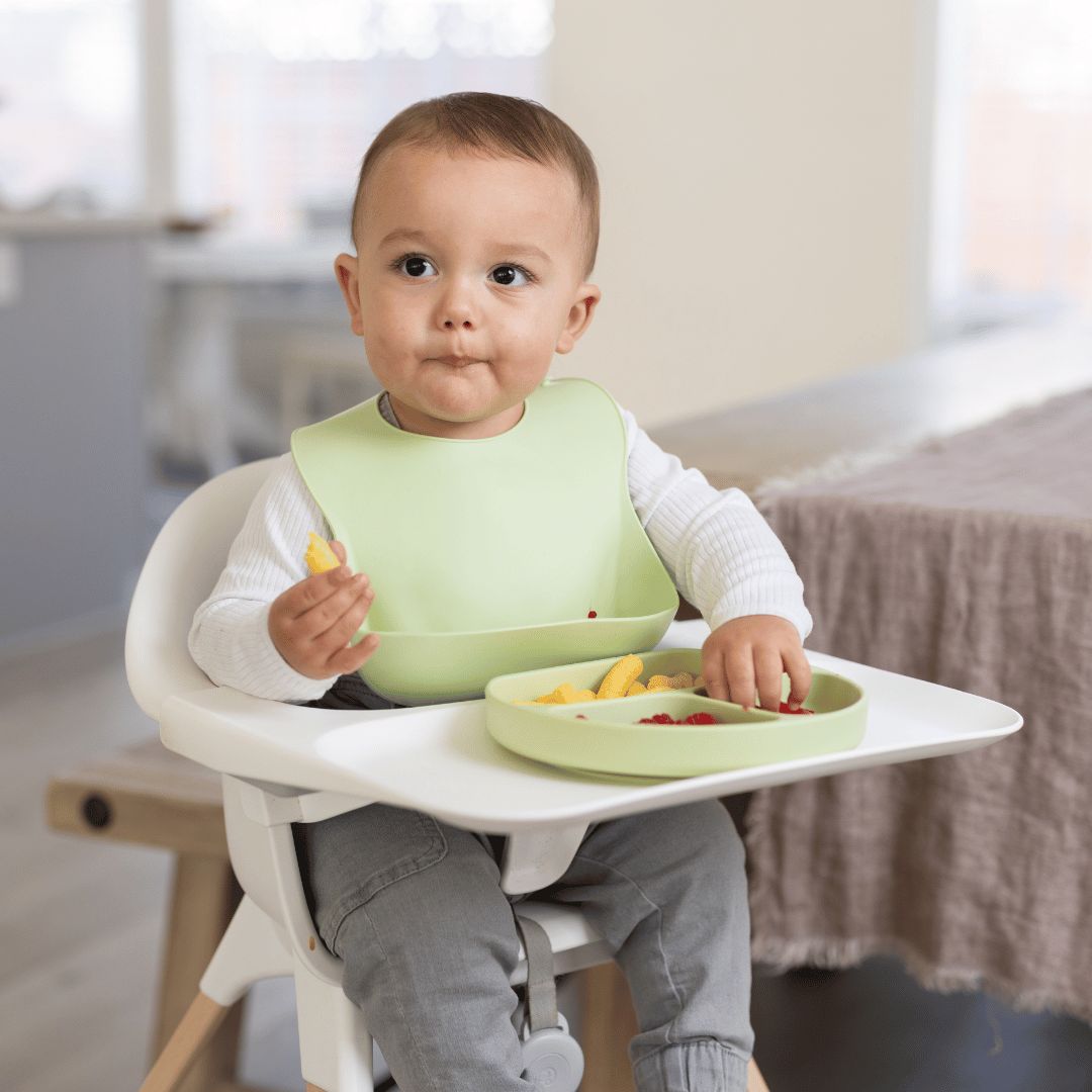 Baby sitting in a high chair enjoying a meal with the Stella Moon silicone suction plate in Spearmint. The baby is wearing a matching silicone bib with a food catcher while self-feeding with a divided plate containing snacks and fresh berries.