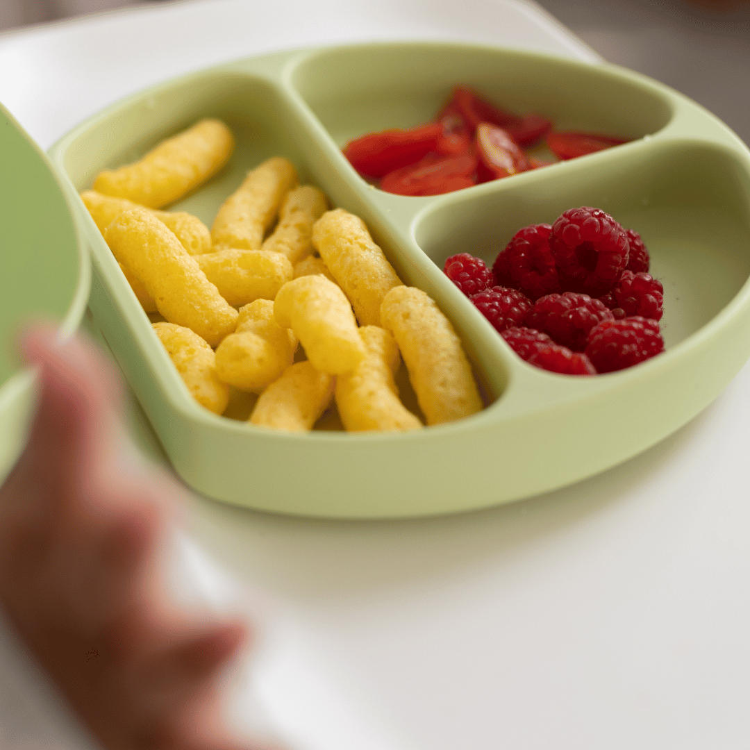 Close-up of the Stella Moon silicone suction plate in Spearmint, filled with raspberries, tomato slices, and puffed snacks. The divided sections keep food separate for easy self-feeding.