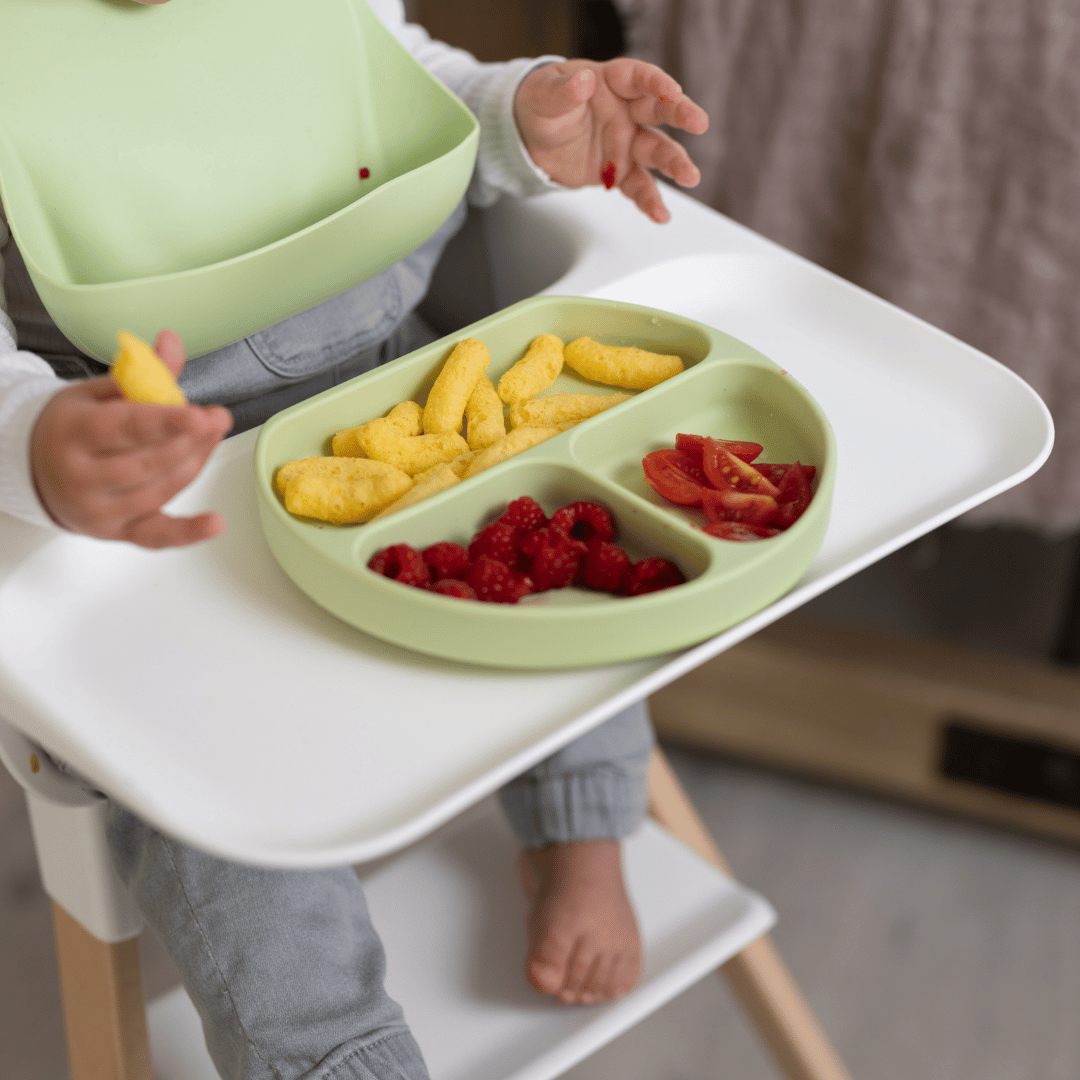 Close-up of the Stella Moon silicone suction plate in Spearmint on a high chair tray. The divided plate contains raspberries, tomato slices, and puffed snacks, designed for easy self-feeding.