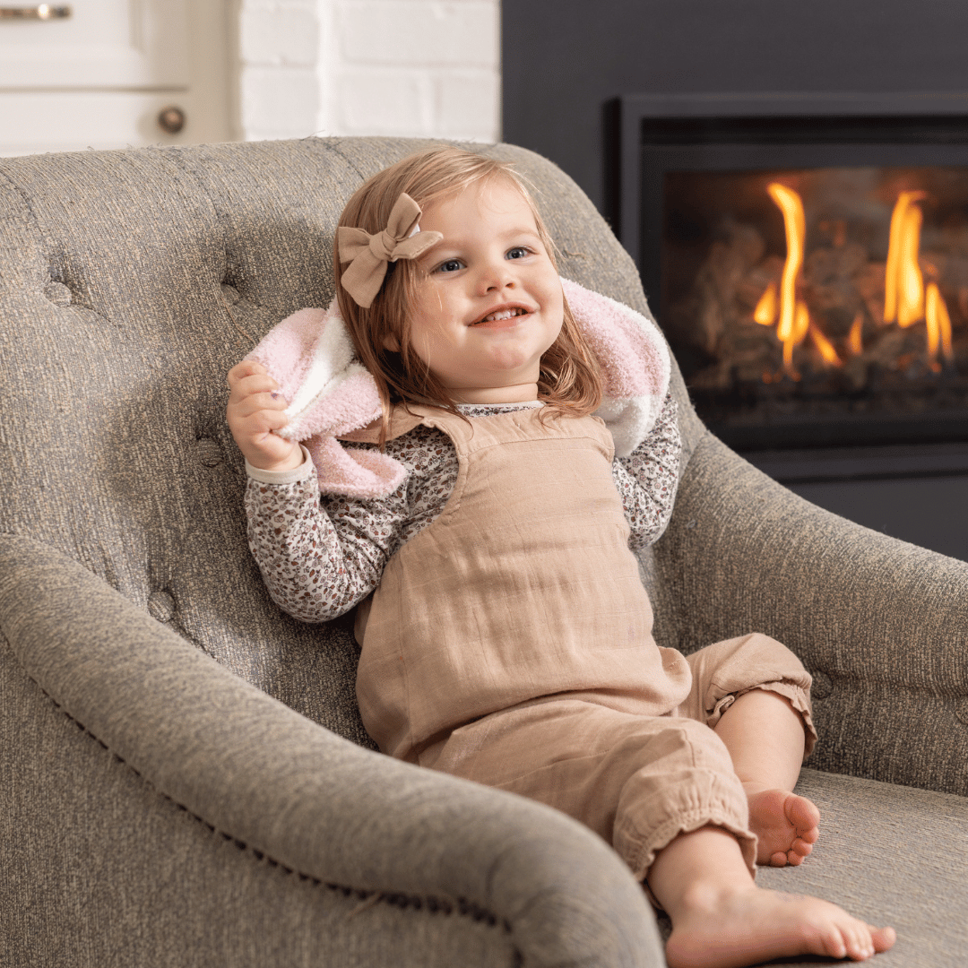 Smiling toddler relaxing in a chair with a soft pink checkered mini baby blanket, adding a touch of comfort and warmth near a cozy fireplace.