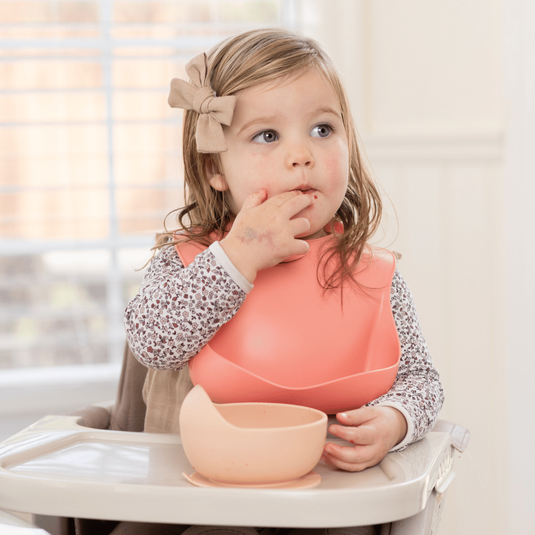 Toddler girl wearing a pink silicone bib, eating a snack with her hands, promoting mess-free mealtime.