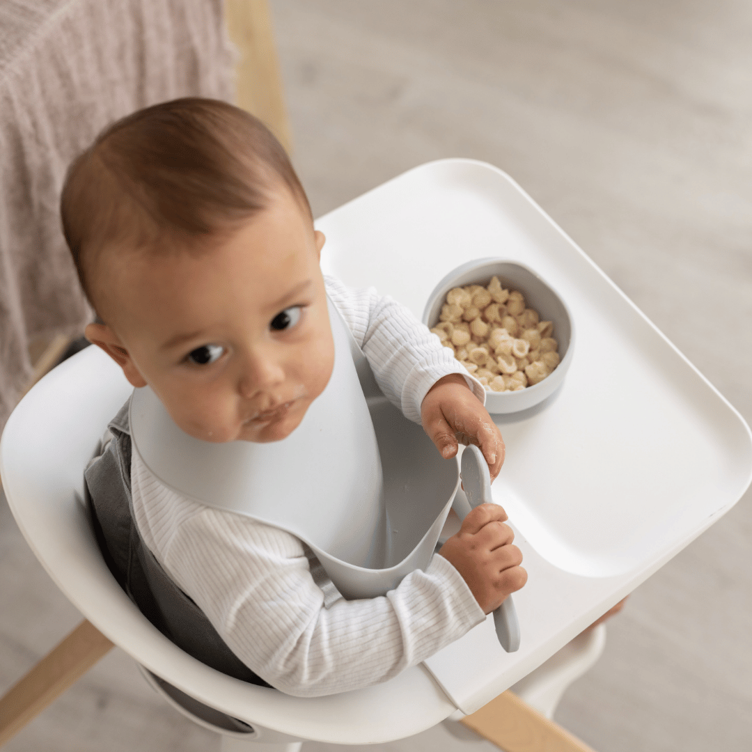 Toddler sitting in a high chair using the Silicone Suction Bowl & Spoon Set in Cloud for self-feeding. BPA-free, non-slip base, perfect for baby-led weaning and easy mealtime.