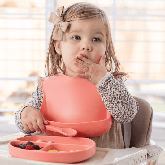 Toddler girl eating with a silicone baby bib in sherbert color, sitting at a table with a bowl of fruits.