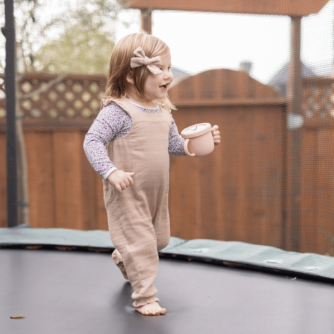 Toddler girl in overalls, playing on a trampoline, holding a blush silicone snack cup.