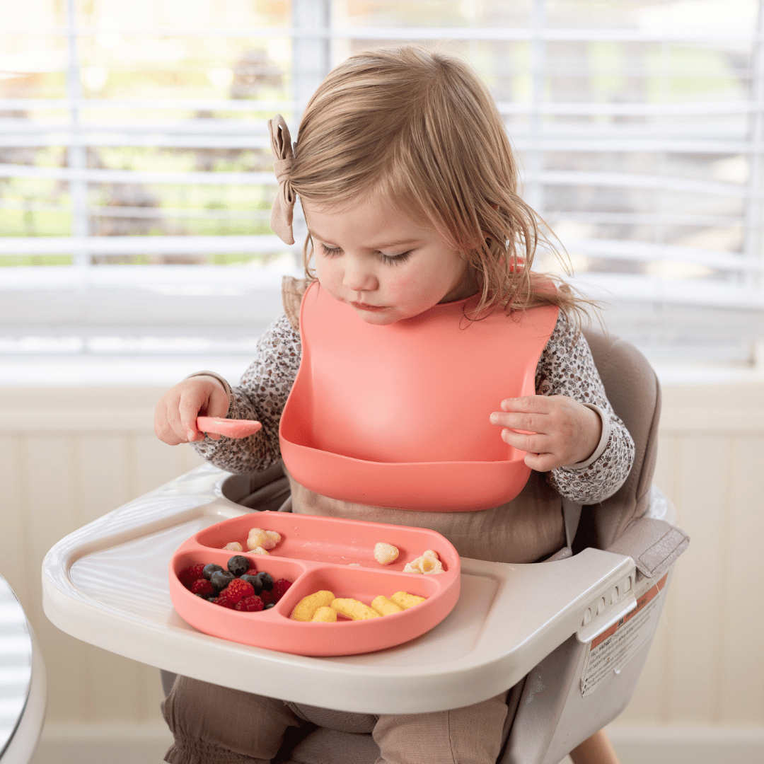 Toddler girl wearing a soft pink silicone bib while exploring her meal in a divided plate, focusing intently on picking up food.