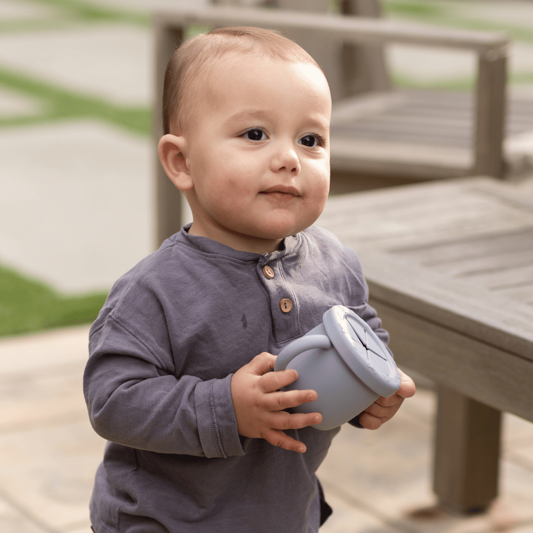  Toddler boy in a grey shirt holding a blue silicone snack cup outdoors.