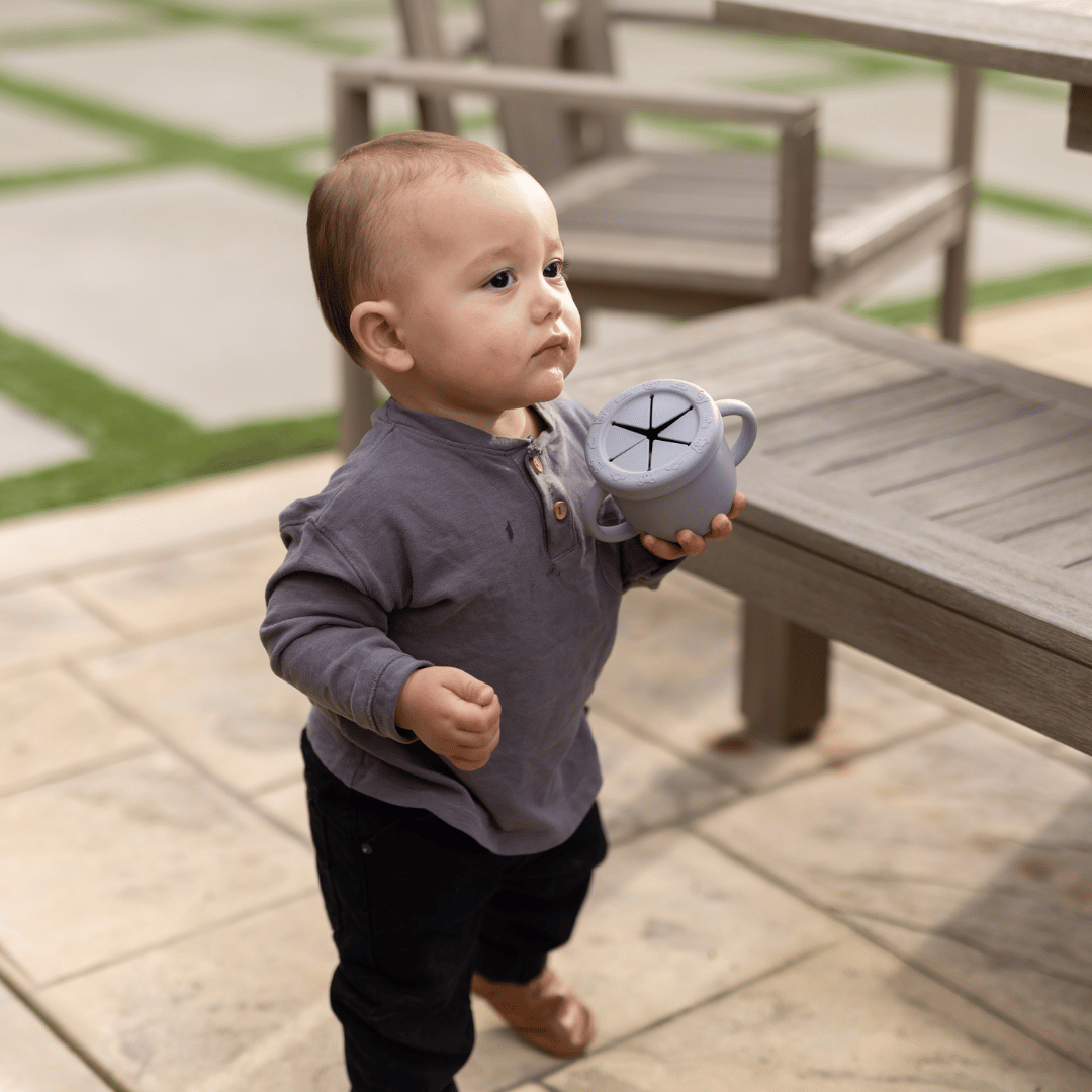 Toddler boy holding a silicone snack cup while standing on a wooden deck, dressed in a grey shirt and black pants.