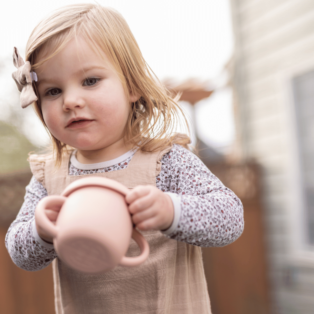 Toddler girl in a beige jumpsuit and floral shirt, holding a pink silicone snack cup, focusing intently on her snack.