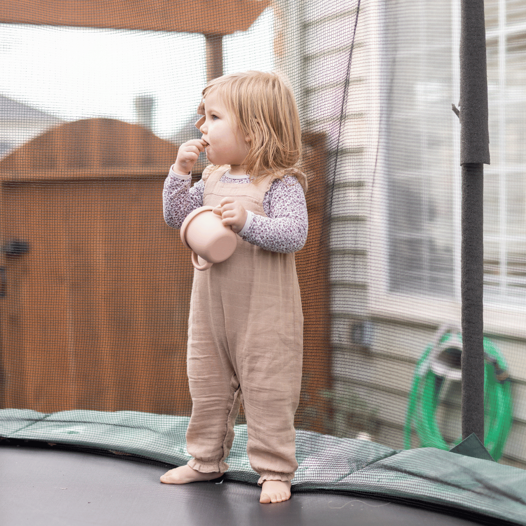 Toddler enjoying a snack on a trampoline, wearing a stylish silicone bib and holding a silicone cup in a natural outdoor setting.