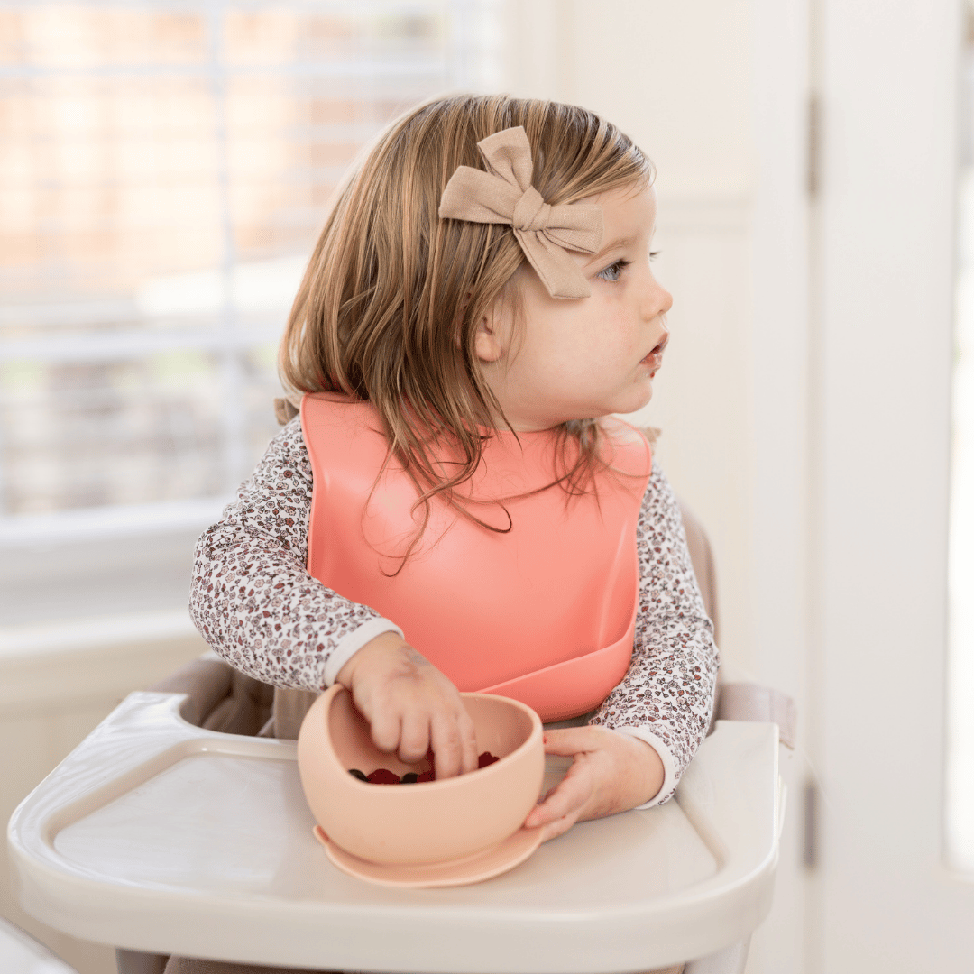  Toddler girl wearing a pink silicone bib, reaching for berries in a matching silicone bowl during mealtime.