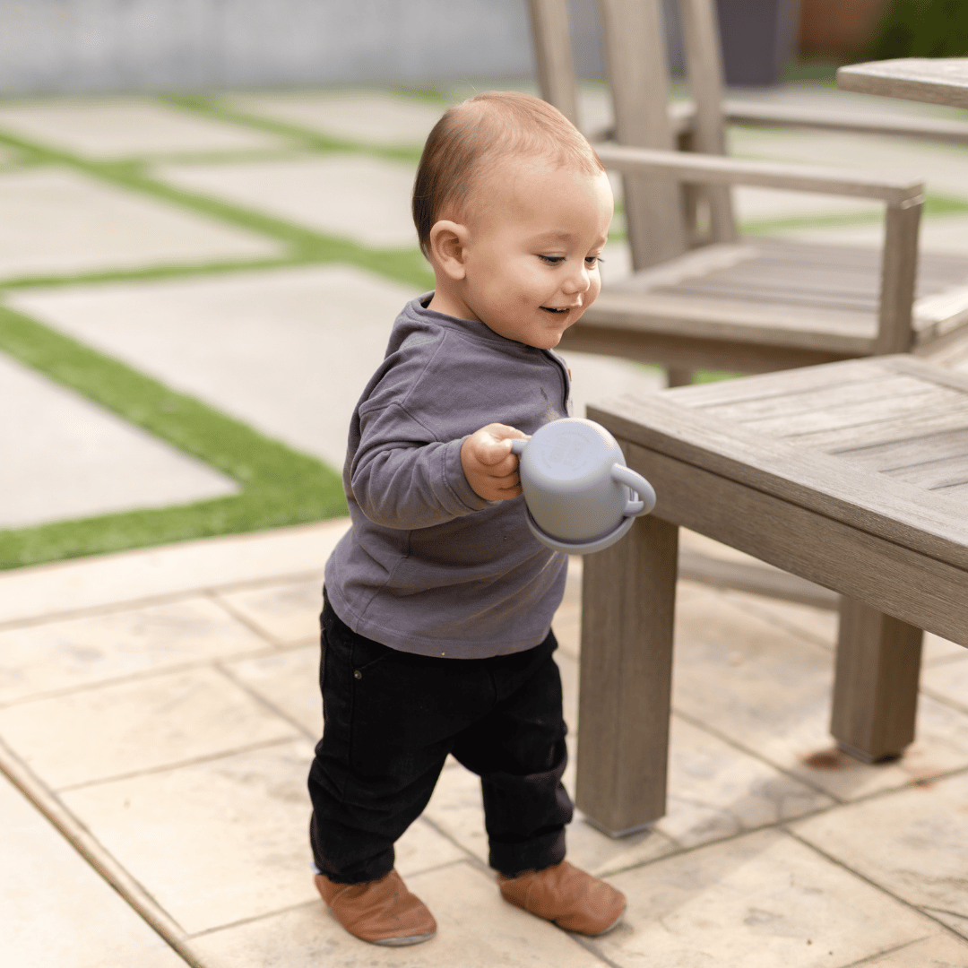 Happy toddler boy in grey shirt and black pants, holding a silicone snack cup while walking outdoors.