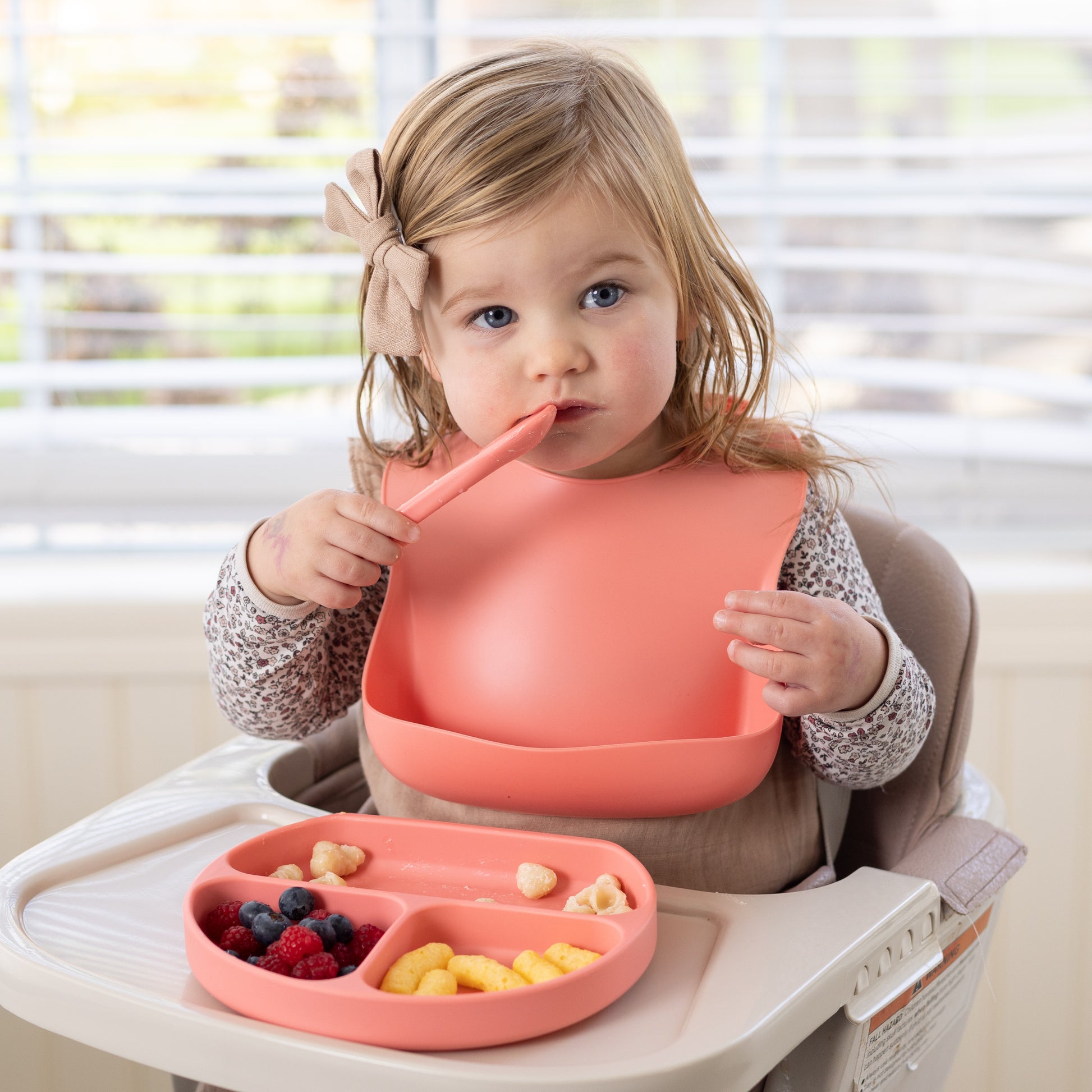 Adorable baby using StellaMoon's Silicone Suction Plate in Sherbert, complemented with a matching bib. The plate's three sections are filled with food, demonstrating its practicality for self-feeding and mess prevention. The BPA-free, non-toxic food-grade silicone material is evident in the plate and bib's vibrant sherbert color.