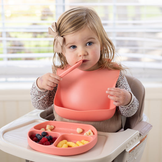 Adorable baby using StellaMoon's Silicone Suction Plate in Sherbert, complemented with a matching bib. The plate's three sections are filled with food, demonstrating its practicality for self-feeding and mess prevention. The BPA-free, non-toxic food-grade silicone material is evident in the plate and bib's vibrant sherbert color.